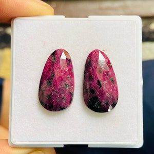 May include: Two teardrop-shaped, faceted gemstones with a deep magenta hue and black inclusions. The gems are displayed in a white plastic case, showcasing their polished surfaces and unique patterns. These stones are likely for jewelry making.