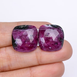 Könnte beinhalten: Zwei kissenförmig geschliffene Edelsteine in tiefem Magenta, mit inneren Adern und Facetten. Die Edelsteine haben schwarze und weiße Einschlüsse an den Rändern. Die Steine werden zwischen Fingern vor weißem Hintergrund gehalten.