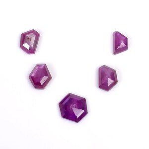 Puede incluir: Cinco cabujones de piedras preciosas de color púrpura en varias formas, incluyendo un hexágono, un diamante y un triángulo. Las piedras preciosas están facetadas y tienen un acabado brillante.