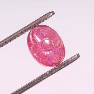 Op de afbeelding: Een gegraveerde roze robijn edelsteen in de vorm van een bloem, vastgehouden door een pincet.