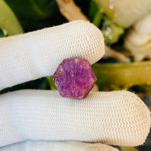 May include: A deep pink, faceted gemstone with a hexagonal shape. The gemstone is held in a white, textured glove.