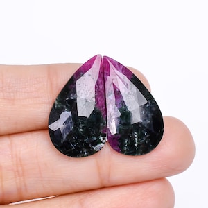 May include: Two heart-shaped gemstones with a faceted cut. The gems have a deep magenta hue at the top, transitioning to a dark green and black base. The stones are displayed against a white background.