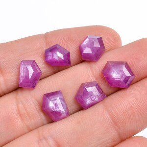 May include: Six pink faceted gemstones with a star-like pattern inside. The gemstones are displayed on a hand.