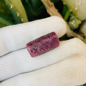 May include: A rectangular, raw ruby gemstone with a deep magenta hue. The stone has a rough, textured surface and is held against a white knitted fabric. The background features green and white foliage.