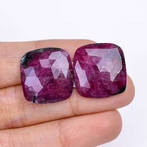 Può includere: Due gemme sfaccettate, taglio a cuscino, di un profondo colore magenta. Le gemme hanno una complessa struttura interna con inclusioni visibili e una superficie lucida. Le pietre sono tenute in mano su sfondo bianco.