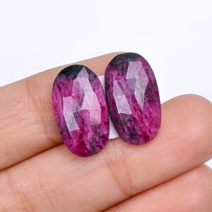 May include: Two faceted, oval-shaped gemstones. The gems are a deep magenta color with darker areas and a polished, reflective surface. The stones are set against a white background.