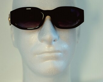 May include: Black sunglasses with a rectangular frame and dark lenses. The sunglasses have a gold accent on the temples. The sunglasses are displayed on a white foam head.