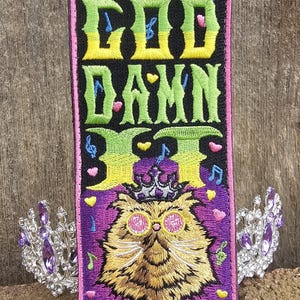 May include: Embroidered patch with the text "GOD DAMN IT" in green, yellow, and blue. The patch features a cat wearing sunglasses and a crown, surrounded by musical notes and hearts. A silver tiara with purple gems is on each side.
