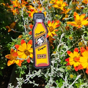 May include: A black and yellow embroidered patch of a bottle of "Deadshot Daiquiri" with a skull and crossbones logo. The bottle is surrounded by yellow flowers with orange tips.
