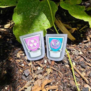 May include: Two shot glass-shaped epoxy resin keychains. One is pink with a white radioactive symbol and the other is blue with a white spiral design.