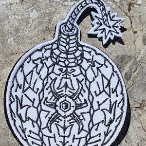 May include: A circular embroidered patch featuring a black and white graphic design of a spherical bomb with a lit fuse. The central motif displays intricate, symmetrical line work resembling a stylized spider or geometric pattern on a white background.
