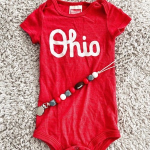 Könnte beinhalten: Roter Baby-Strampler mit weißem "Ohio"-Schriftzug und einem grauen, weißen und roten Silikon-Beißring mit einem Fußball-Anhänger.