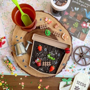 May include: A miniature gardening set with a brown pot, green shovel, and a container of garden soil. The set includes a watering can, seed packets, and decorative elements like carrots, a ladybug, and a caterpillar. The set is arranged on a wooden slice.