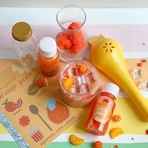 May include: A set of orange juice making supplies, including a glass pitcher, a yellow citrus squeezer, a glass bottle with a gold lid, a glass bottle with a white lid, a glass jar with a lid, a glass jar with orange and red sprinkles, and a card with instructions for making orange juice. The card says "Fresh squeezed orange juice" and has illustrations of oranges, a squeezer, and a pitcher.
