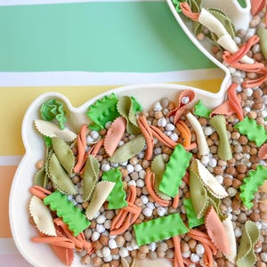 May include: A white bunny-shaped plate filled with colorful pasta and chickpeas. The pasta includes green, orange, and white shapes, resembling carrots and leaves. The background features pastel stripes of yellow, green, and blue.