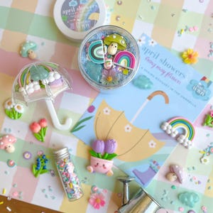May include: A collection of colorful spring-themed decorations, including miniature rainbows, mushrooms, flowers, and an umbrella. The items are arranged on a pastel checkered background, with a jar of sprinkles and a container with the text "april showers".