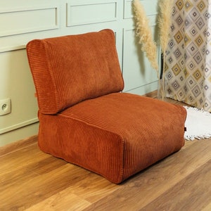 Op de afbeelding: Een bruine corduroy vloercushion met een bijpassende rugleuning. De cushion staat op een houten vloer in een kamer met een lichtgroene muur en een schilderij aan de muur.