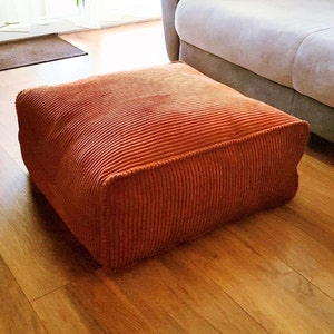 May include: A large, orange corduroy floor cushion. The cushion is square shaped and has a thick, plush texture.