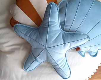 Puede incluir: Tres almohadas de felpa con forma de estrella de mar, concha y vieira. La estrella de mar y la concha son de color azul claro, y la vieira es de color blanco y marrón.