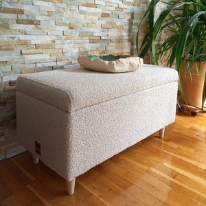 May include: A white, fluffy storage ottoman with a wooden base. The ottoman is sitting on a wooden floor in front of a stone wall.