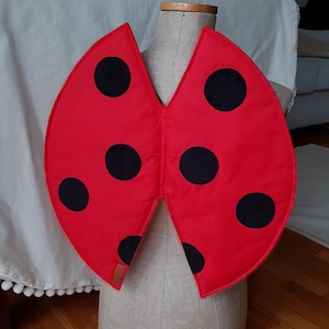 May include: Red ladybug wings costume with black spots. The wings are made of fabric and are attached to a piece of elastic.