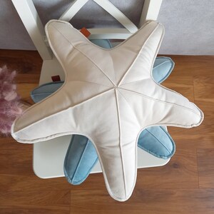 Può includere: Un cuscino a forma di stella marina color crema è appoggiato sopra un cuscino a forma di stella marina azzurra. Entrambi i cuscini sono su una sedia bianca. I cuscini hanno un aspetto morbido e soffice, con dettagli cuciti. I cuscini sono decorativi.