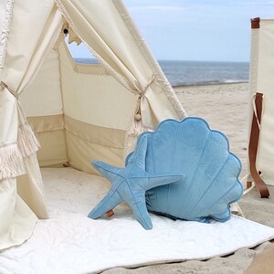 May include: A light blue velvet starfish and seashell pillow set. The pillows are placed inside a white teepee tent on a white rug. The teepee is set up on a sandy beach.