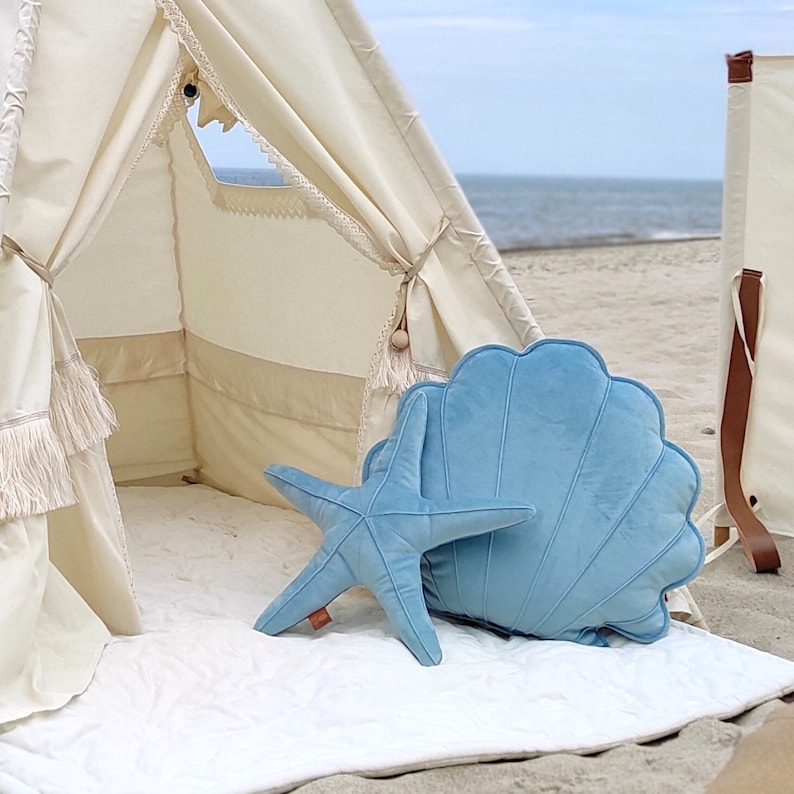 May include: A light blue velvet starfish and seashell pillow set. The pillows are placed inside a white teepee tent on a white rug. The teepee is set up on a sandy beach.