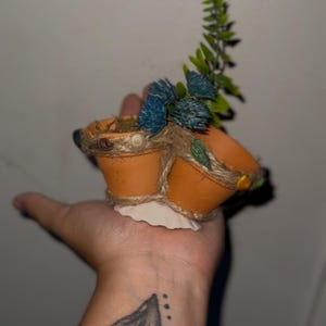 May include: A small, decorative terracotta pot with three sections, adorned with twine, seashells, and faux greenery. The pot is filled with artificial plants, including blue and green accents. A unique, handcrafted item.