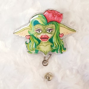 Op de afbeelding: Een groene en roze glitter badgehouder met een cartoonfiguur met groen haar, rode lippen en een roze bloem.
