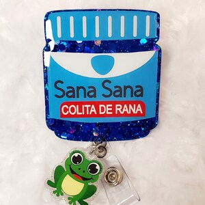 Könnte beinhalten: Ein blau glitzernder Ausweis-Rollenhalter in Form einer Medikamentenflasche mit den Worten "Sana Sana COLITA DE RANA" in Weiß und Rot. Ein grüner Frosch-Anhänger ist befestigt.