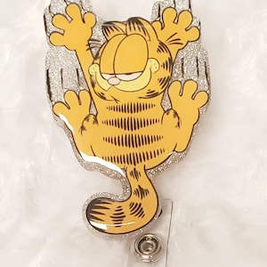 Peut inclure: Un porte-badge en paillettes argentées avec une image de dessin animé de Garfield le chat suspendu à celui-ci. Garfield est orange et noir et est représenté les bras levés en l'air.