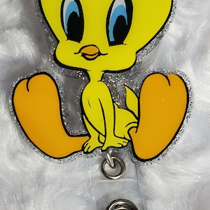 Op de afbeelding: Een gele cartoonvogel met grote ogen en een kleine snavel, zittend op een glinsterende, transparante achtergrond. De vogel is bevestigd aan een transparante plastic badgehouder met een zilveren metalen clip.