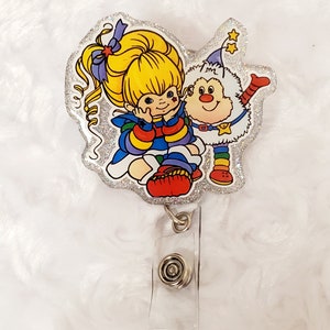 Puede incluir: Un portagafete de purpurina plateada con una imagen de dibujos animados de una niña con rayas arcoíris y una criatura peluda blanca con una cola arcoíris. La niña tiene el pelo rubio y un lazo azul. La criatura tiene la nariz roja y un sombrero azul con estrellas.