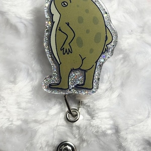 May include: A green frog-shaped badge reel with a silver glitter border. The frog has a light green body with darker green spots and a cartoonish design. The reel has a clear plastic strap and a metal clip.
