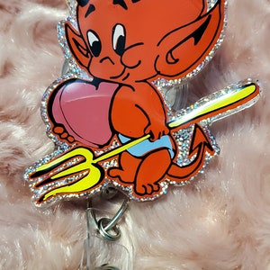 Puede incluir: Un carrete de insignia con temática de diablo rojo con un diablo de dibujos animados sosteniendo un corazón rosa y un tridente amarillo. El carrete tiene un borde brillante y un clip de plástico transparente.