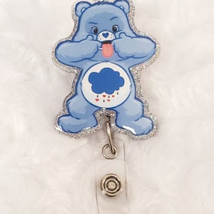 Peut inclure: Un ours en peluche bleu pailleté avec un nuage et des gouttes de pluie sur le ventre. L'ours sort sa langue et fait une grimace amusante. L'ours est attaché à un porte-badge rétractable en plastique transparent avec un clip en métal argenté.