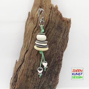 Può includere: Un portachiavi verde e argento con una pila di pietre lisce grigie, bianche e gialle. Due ciondoli a forma di pesce argentati pendono dalla parte inferiore del portachiavi. Il portachiavi è esposto su un pezzo di legno galleggiante.