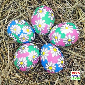 Pode incluir: Cinco ovos de Páscoa pintados à mão com um padrão de margaridas em tons de rosa, verde e azul, sobre um fundo de palha. Cada ovo apresenta margaridas brancas com centros amarelos. Os ovos estão dispostos em círculo.