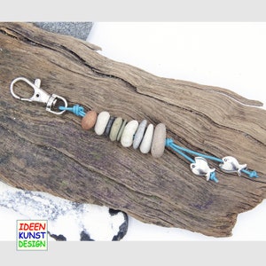 Può includere: Un portachiavi blu e argento con una serie di pietre lisce grigie e due ciondoli a forma di pesce argentati. Il portachiavi è su una superficie di legno.
