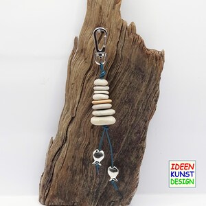 Può includere: Un portachiavi in pietra bianca e beige con un moschettone argentato e due ciondoli a forma di pesce argentati appesi a un cordoncino blu. Il portachiavi è appeso a un pezzo di legno di deriva.