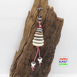 Può includere: Un portachiavi rosa e argento con una pila di pietre lisce grigie e due ciondoli a forma di pesce argentati. Il portachiavi è appeso a un pezzo di legno di deriva.