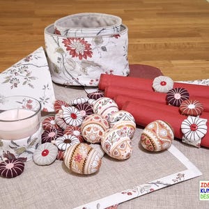 Peut inclure: Une table dressée avec une nappe à motifs floraux, des serviettes rouges avec des anneaux en tissu et une variété d'œufs de Pâques peints. La table est également décorée d'une bougie dans un bocal en verre et d'un panier de rangement à motifs floraux.