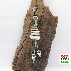Può includere: Un portachiavi argentato con una pila di pietre grigie, bianche e marroni. Le pietre sono legate insieme da un cordoncino verde e hanno due ciondoli a forma di pesce argentati che pendono dal fondo.