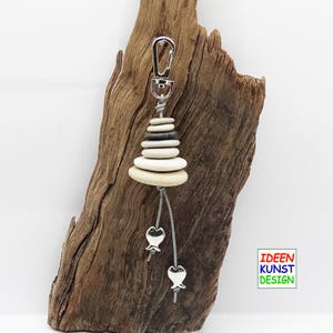 Può includere: Un portachiavi a forma di pila di pietre bianche e grigie con un anello portachiavi argentato e due ciondoli a forma di pesce argentati appesi a cordini grigi. Il portachiavi è appeso a un pezzo di legno di deriva. Il testo "IDEEN KUNST DESIGN" è visibile nell'angolo in basso a destra dell'immagine.