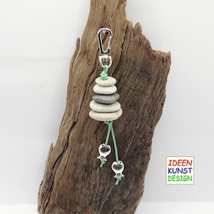 Può includere: Un portachiavi argentato con una pila di pietre grigie e bianche e due ciondoli a forma di pesce argentati appesi a un cordoncino verde. Il testo "IDEEN KUNST DESIGN" è visibile nell'angolo in basso a destra dell'immagine.