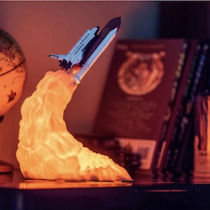 May include: A white space shuttle shaped night light with a glowing orange cloud base, resembling a rocket launch. The night light is on a dark wooden surface.