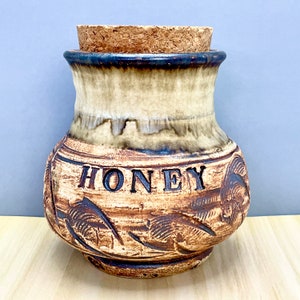 May include: A ceramic honey pot with a cork lid. The pot is decorated with a brown and white glaze and has the word "HONEY" written on the front. The pot has a rustic, hand-crafted look.