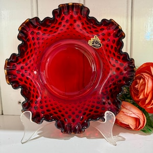 May include: A bright red Fenton hobnail glass plate with a frilled edge. The plate features a circular centre and is supported by a clear stand. The plate has a hobnail pattern and a Fenton sticker.