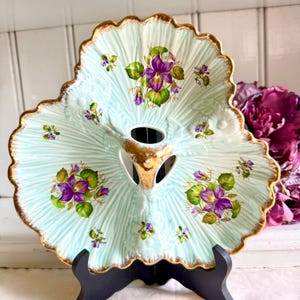 May include: A vintage, light blue, leaf-shaped serving dish with a gold rim and accents. The dish features a floral design with purple violets and green leaves. The dish is supported by a black stand.